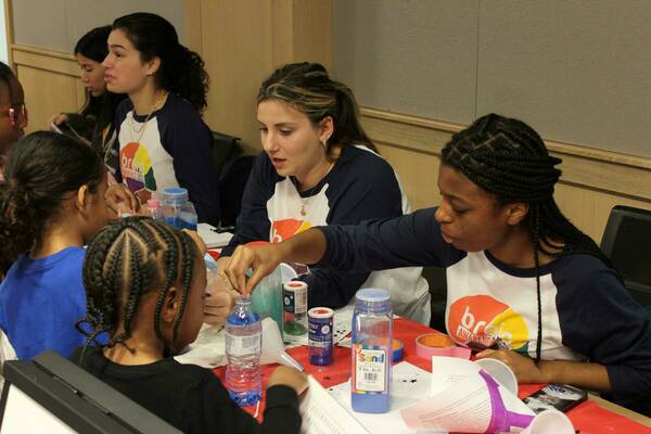 Notre Dame students help children make glitter jars at a Brain Awareness Week event. The students wear navy and white shirts with the Brain Awareness logo.  On the table are bottles of glitter, colored sand, funnels, and water bottles.