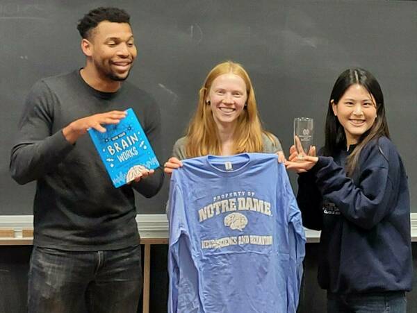 Three individuals stand before a chalkboard. One holds a book titled "How Your Brain Works," another displays a light blue, long-sleeved Notre Dame Neuroscience and Behavior t-shirt, and the third holds a clear glass award.