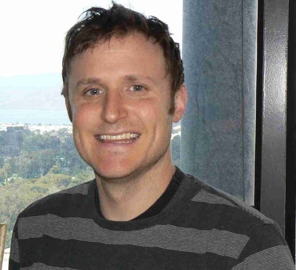 Smiling man with short brown hair and blue eyes, wearing a dark gray striped shirt. Cityscape and water visible in blurred background.