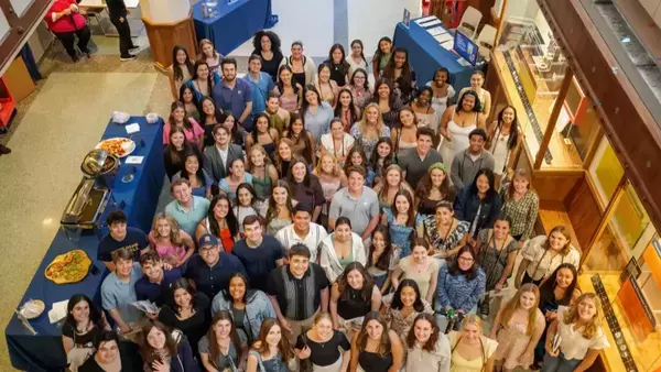Overhead view of many diverse Notre Dame students smiling, gathered around blue-clothed food tables in a campus building.